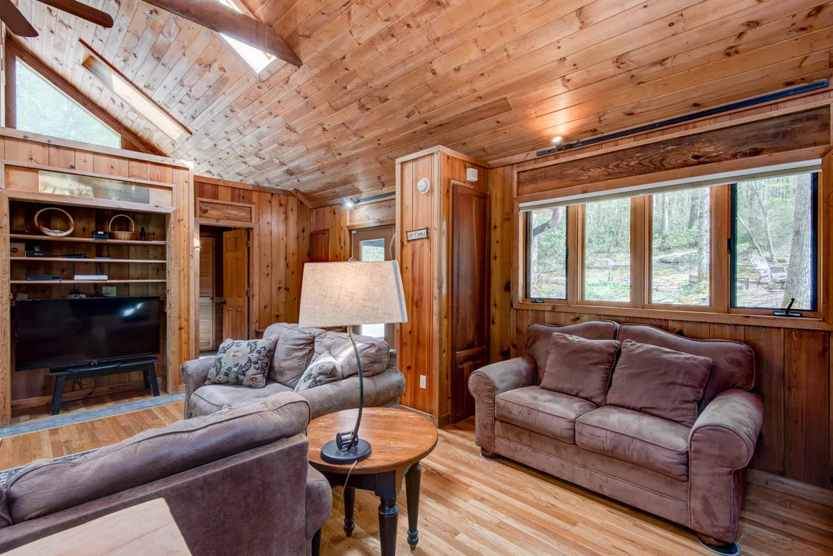 A spacious living room with warm wooden ceilings and beams, large windows that frame lush woodland views, plush comfortable sofas, and a table lamp. The room feels open and inviting, with natural light pouring in to highlight the rustic details and cozy layout ideal for gatherings.