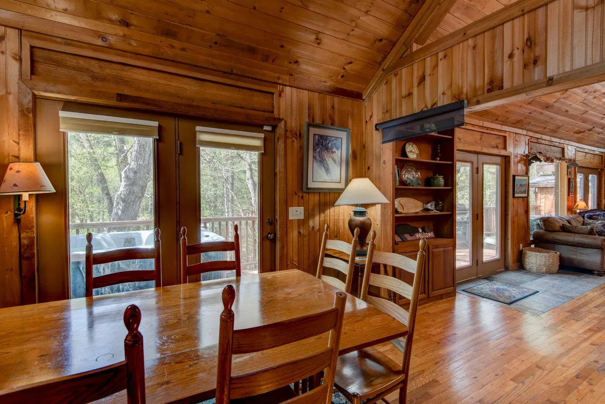 A welcoming dining area featuring a sturdy wooden table, exposed beams overhead, large windows letting in natural light, a lamp, and a well-stocked bookshelf. The space feels perfect for shared meals, board games, or intimate gatherings, all set against a backdrop of forest views.