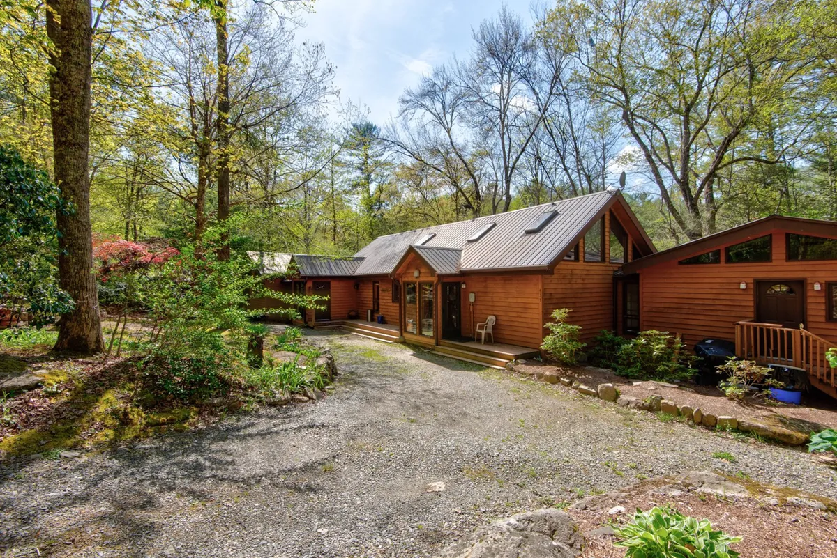 A wide exterior view of the wooden cabin surrounded by a landscaped garden and gravel driveway, nestled among tall trees. The inviting porch provides an ideal place to enjoy quiet mornings or evenings, set against a backdrop of natural beauty.
