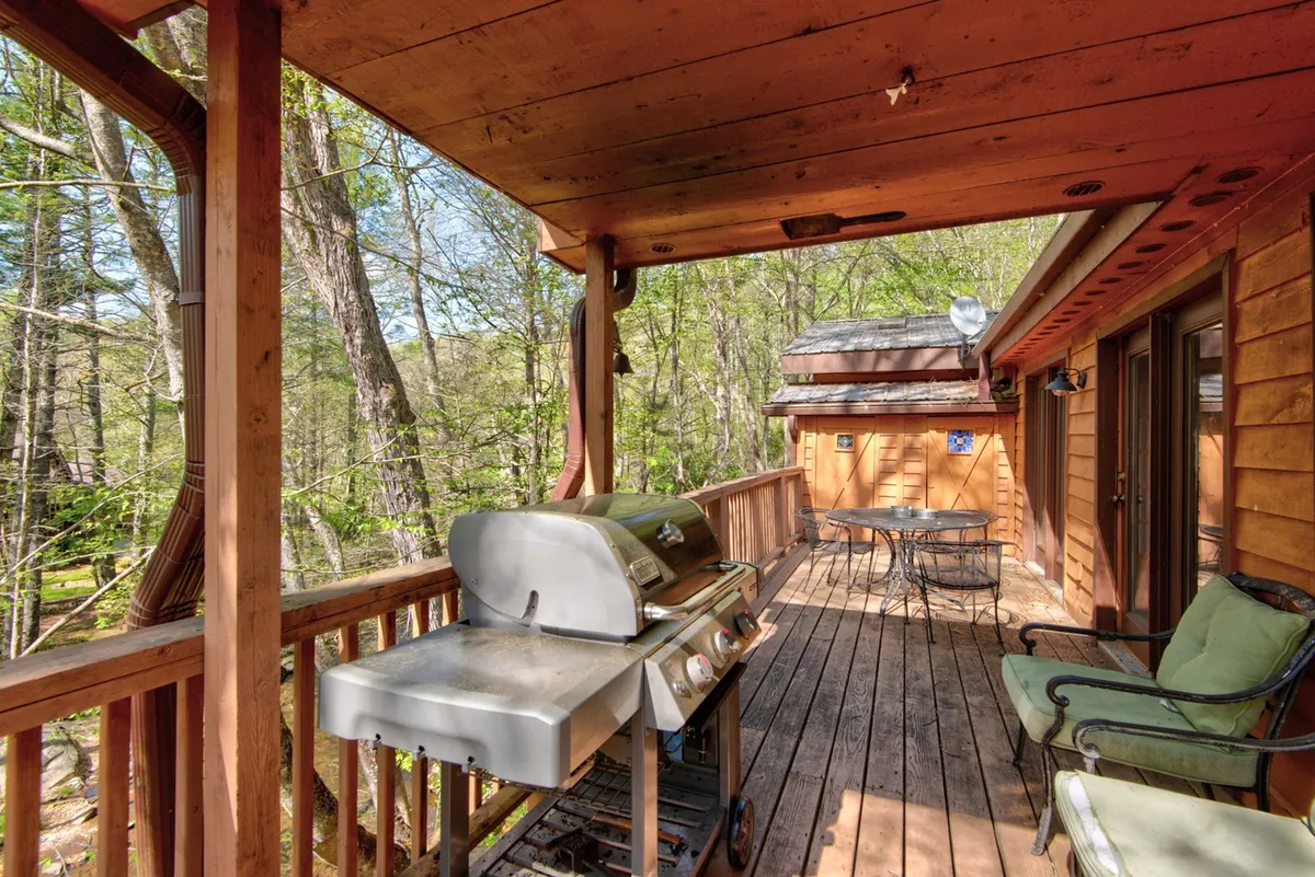 A wooden deck with a dining table, seating area, and grill, framed by a wooden railing and overlooking lush greenery. The setup is ideal for alfresco dining, social gatherings, or evening relaxation under the stars.
