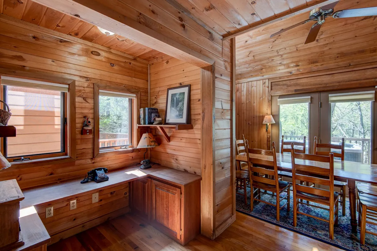 A warm, wood-paneled space featuring a dining table, windows bringing in daylight, and a desk area thoughtfully positioned for work or reading. The setting feels inviting and multifunctional, suited for both productivity and quiet moments.