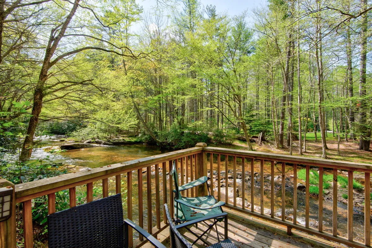A wooden deck overlooks a flowing creek and green trees, with comfortable chairs arranged for relaxation—an inviting spot for morning coffee or group board games outdoors.