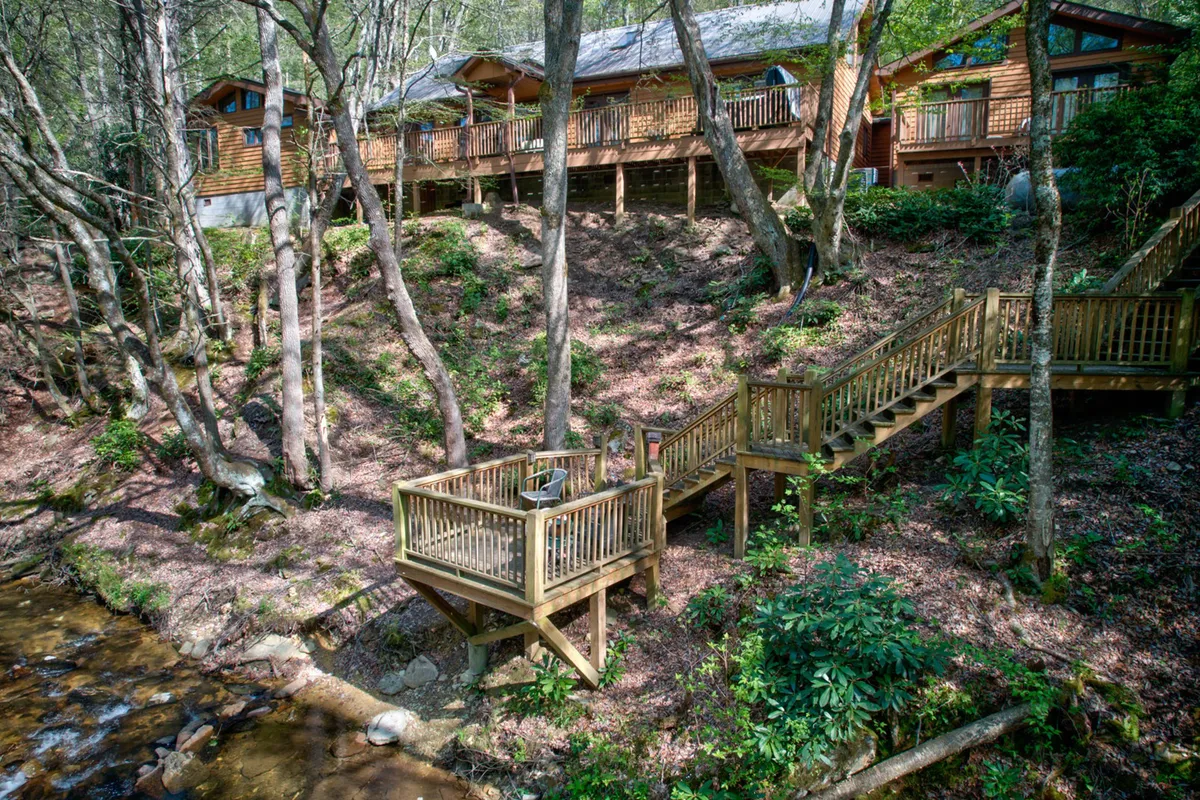 A spacious wooden deck attached to a log cabin-style home, overlooking a stream and surrounded by lush trees. Comfortable seating creates an inviting spot for morning coffee, evening gatherings, or simply soaking in nature after exploring the region.