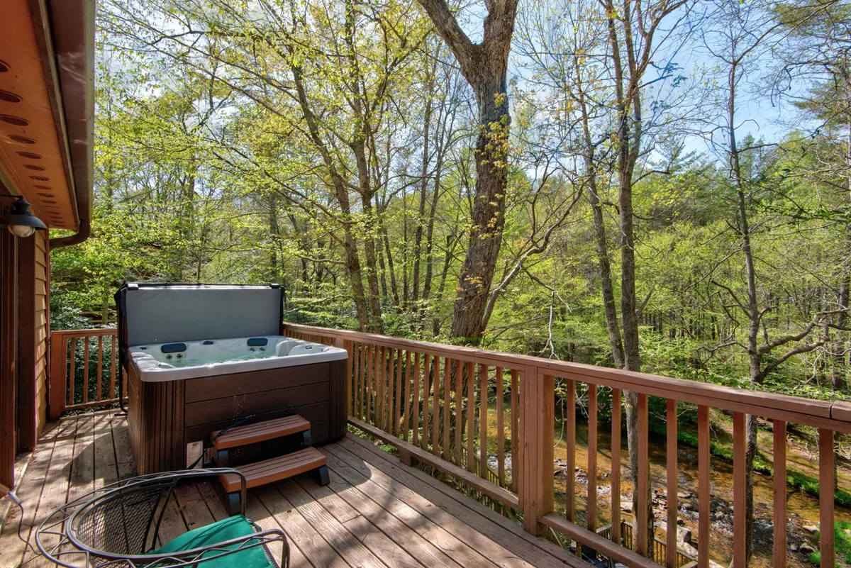 A serene exterior view of a private wooden deck nestled among trees, featuring a hot tub overlooking a peaceful stream. The setting is surrounded by nature, offering a secluded and relaxing atmosphere perfect for unwinding after days spent exploring local mountain towns.