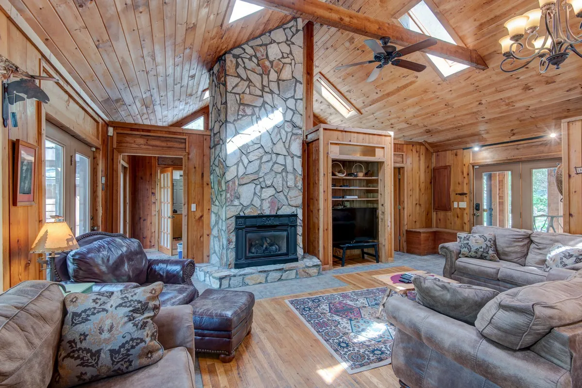 A warm and inviting living room with a prominent stone fireplace, wooden ceiling, large windows, and comfortable seating. The natural light and cozy atmosphere evoke the perfect winter retreat, where guests can gather after snowy adventures or relax by the fire.