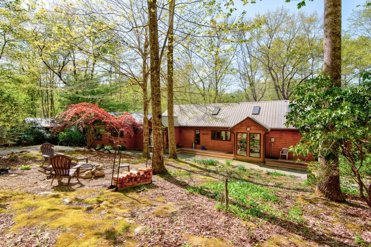 A cozy outdoor seating area surrounding a firepit, set in a landscaped garden beside a wooden house. The image shows stacked firewood, mature trees, and autumn foliage, emphasizing warmth and togetherness.