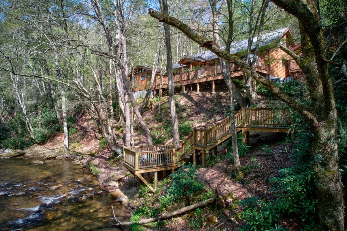 This exterior shot captures the wooden house nestled among trees with a stream and spacious decking, highlighting the property's natural setting and inviting outdoor gathering spaces.