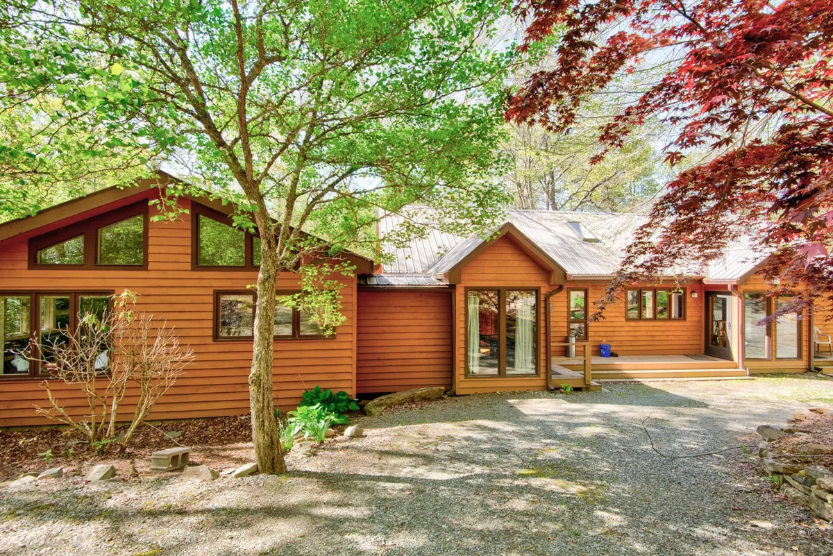 The image showcases a charming wooden cabin with large windows, nestled in a lush, landscaped garden. A gravel pathway leads up to the inviting porch, surrounded by mature trees and greenery, creating a tranquil, private setting perfect for relaxation. The exterior hints at opportunities for enjoying sunny mornings on the porch or gathering in the landscaped yard.