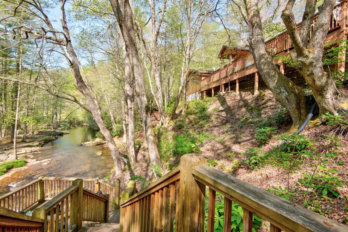 An exterior shot showing wooden stairs leading down to a flowing creek, surrounded by trees. The view from the balcony overlooks the peaceful water and forest, hinting at the nearby hiking trails, riverside relaxation, and opportunities for bird watching and wildlife spotting.
