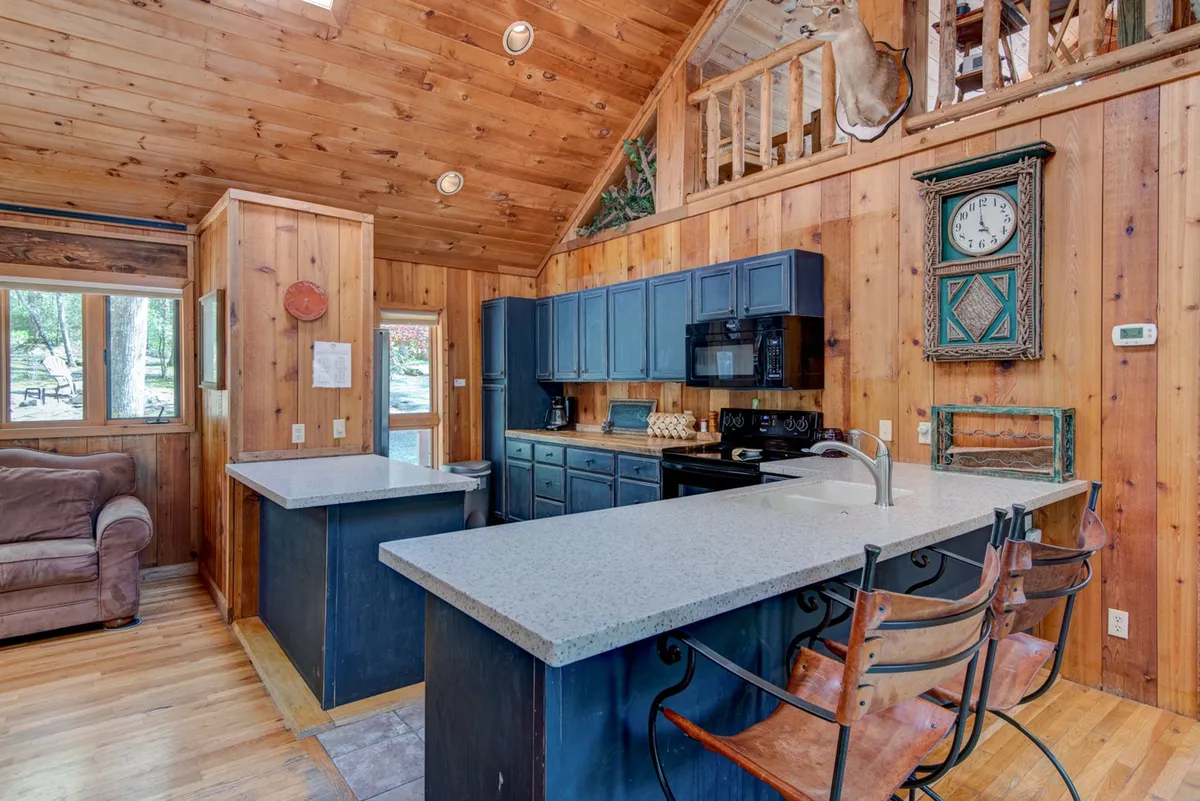 A view of the open kitchen area with a central island, wooden cabinetry, a large window looking out to nature, and a comfortable sofa nearby. The seamless blend of kitchen and living space supports both cooking and socializing, capturing the essence of open-plan living.