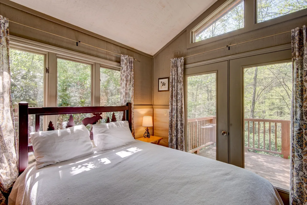 A bright bedroom with a decorative headboard, large windows filling the room with natural light, and a view out to a wooden deck. The bed is neatly made, and the room feels airy and inviting, suggesting a restful retreat with direct access to the outdoors.
