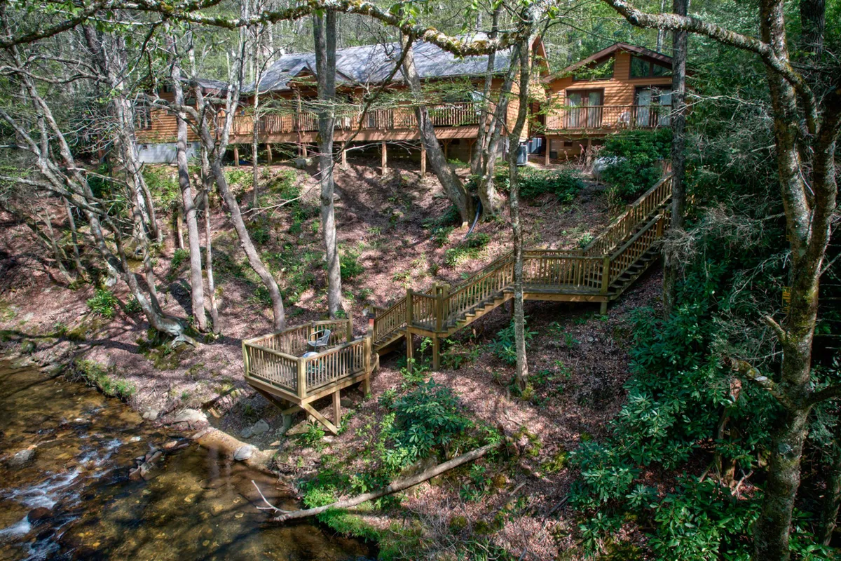 This image shows a tranquil wooden deck overlooking a flowing creek, surrounded by lush trees and featuring the cabin structure in the background. The setting evokes a deep sense of immersion in nature, with the water, forest, and rustic architecture all visible in one frame.